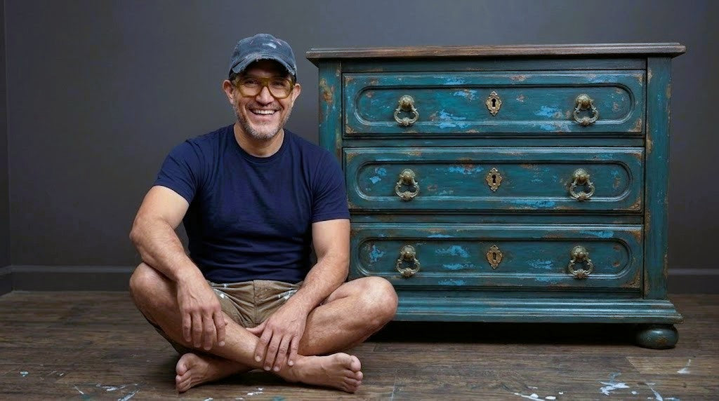 A smiling man, wearing yellow-rimmed glasses, a navy t-shirt, and a weathered baseball cap, sits barefoot with legs crossed on a paint-splattered wooden floor, gesturing toward an antique, distressed teal-blue wooden dresser with detailed gold hardware